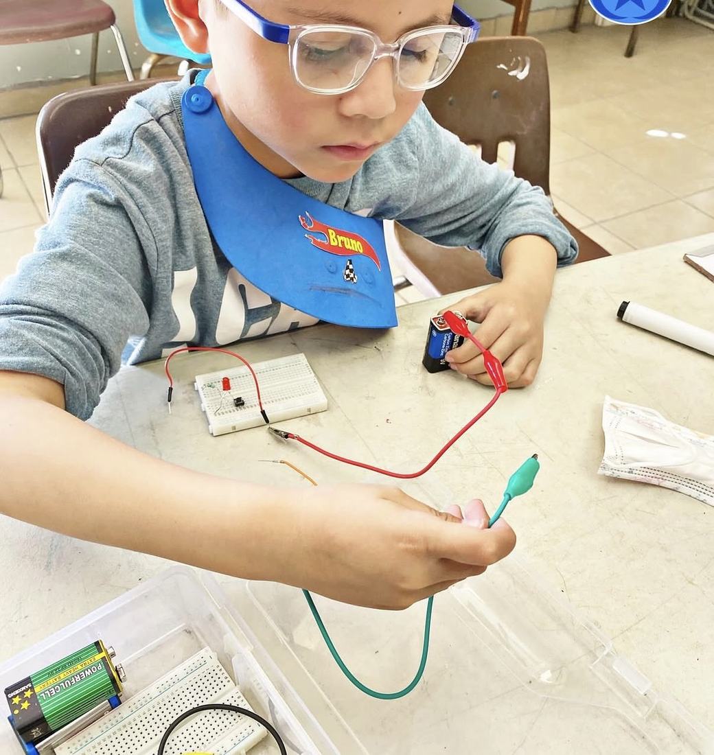 Niño de primaria armando su primer circuito con una pila, cables y un foco en un taller de CultivaTec