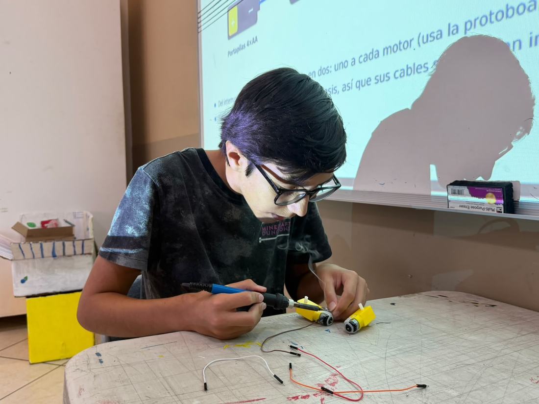 Alumno de secundaria arma un robot móvil con protoboard y motores durante un taller de CultivaTec, con la clase proyectada al fondo